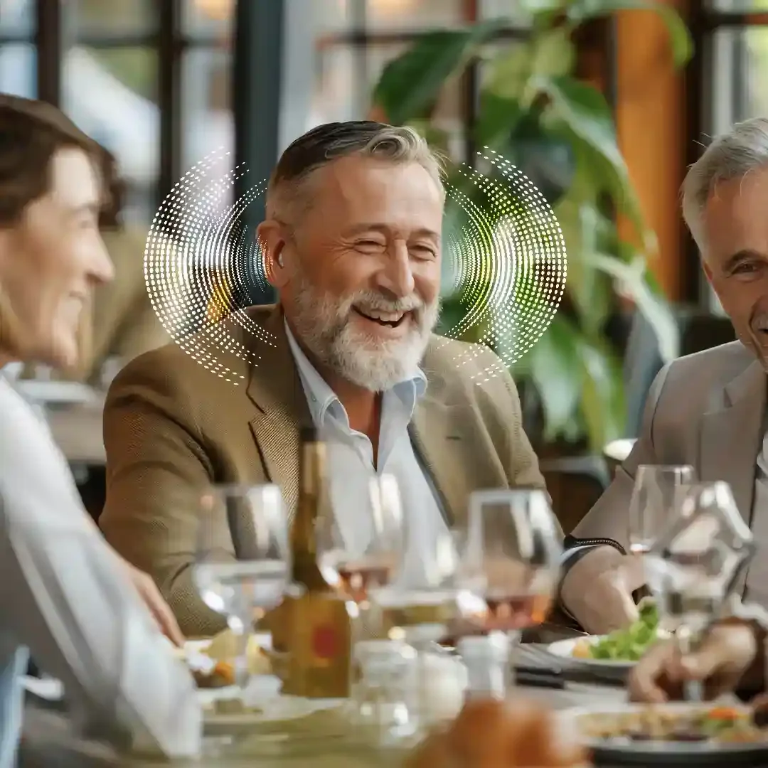 Older man laughing with family at a restaurant table while wearing a hearing aid